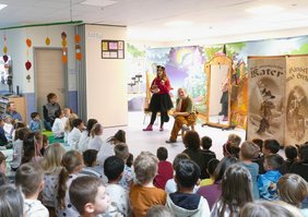 Zahlreiche Kinder sitzen auf dem Boden der Kita Entdeckerland und verfolgen die Aufführung der „Opernretter“. Vor einer bunten Märchenkulisse stehen zwei Darstellerinnen in unterschiedlichen Kostümen. Rechts sind große Bühnenbilder mit den Aufschriften „Der gestiefelte Kater“ und „Hänsel und Gretel“ zu sehen.