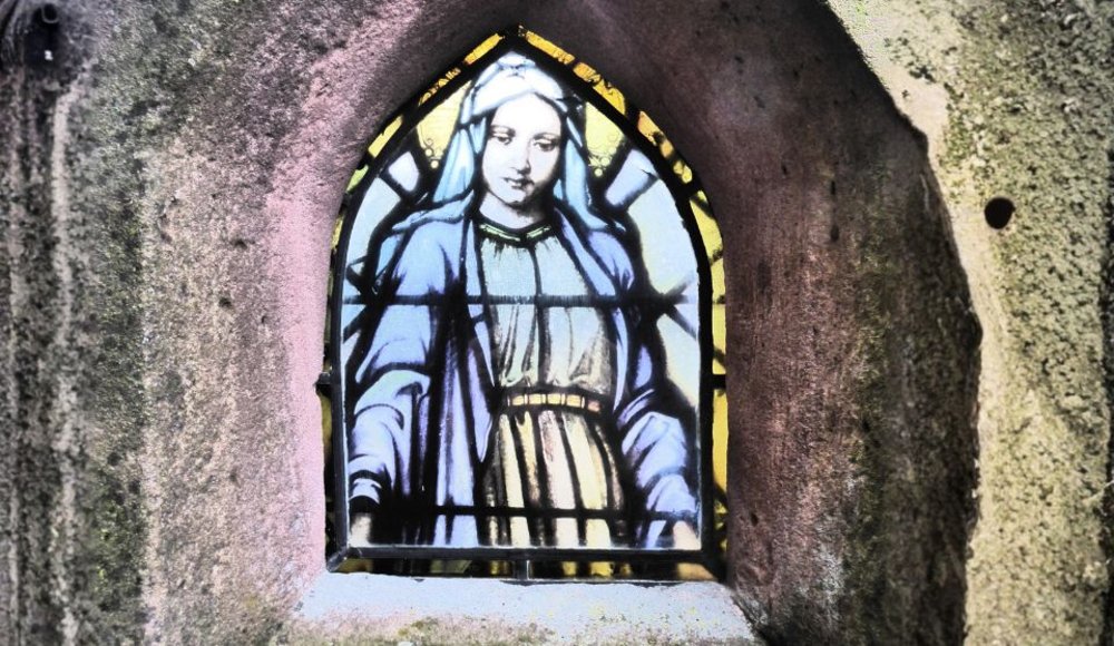Nahaufnahme eines spitzbogenförmigen Buntglasfensters in einer steinernen Einfassung. Das Fenster zeigt eine weibliche, religiös anmutende Figur in einem hellen Gewand und blauem Umhang.