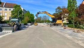 Breite Straßenansicht der Saarlandstraße während Bauarbeiten an einem sonnigen Tag: In der Mitte eine aufgerissene Fahrbahn, rechts ein großer gelber Bagger, der mit seinem Arm über einen Haufen Schotter greift. Links steht ein Radlader mit großer Schaufel. Im Hintergrund sind Absperrungen und ein kleines gelbes Fahrzeug zu sehen. Die Straße wird von mehrstöckigen Wohnhäusern und grünen Bäumen gesäumt, der Himmel ist klar und blau.
