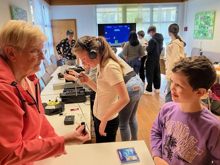 In einem hellen Raum stehen Kinder und ältere Frauen gemeinsam an einem Tisch mit verschiedenen Geräten. Eine Seniorin zeigt einem Jungen ein kleines technisches Gerät, während ein Mädchen mit Kopfhörern einen Kassettenrekorder ausprobiert. Im Hintergrund spielen mehrere Kinder an einem Bildschirm ein klassisches Videospiel.