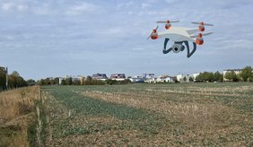 Blick über ein weitläufiges Feld am Stadtrand unter bewölktem Himmel. Im Hintergrund stehen Wohn- und Gewerbegebäude, ebenso das Neue Rathaus. In den Himmel ist die Illustration einer Drohne mit Kamera eingefügt.