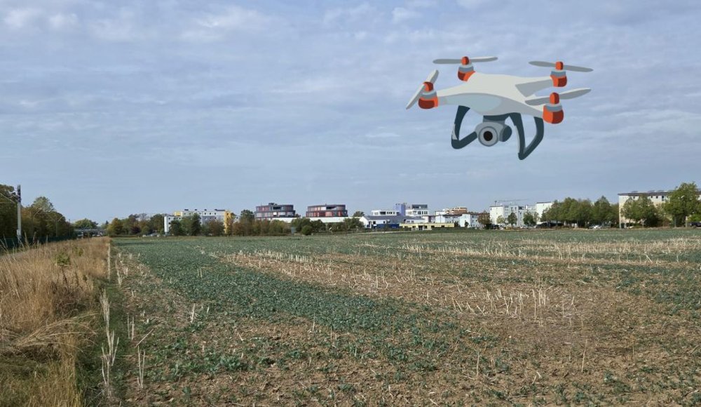 Blick über ein weitläufiges Feld am Stadtrand unter bewölktem Himmel. Im Hintergrund stehen Wohn- und Gewerbegebäude, ebenso das Neue Rathaus. In den Himmel ist die Illustration einer Drohne mit Kamera eingefügt.