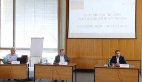 Pressekonferenz vom 28.03.2022 ©Stadt Viernheim Drei Männer sitzen hinter drei Tischen im Hintergrund Leinwand mit Präsentation Unterbringung von Flüchtlingen in Viernheim