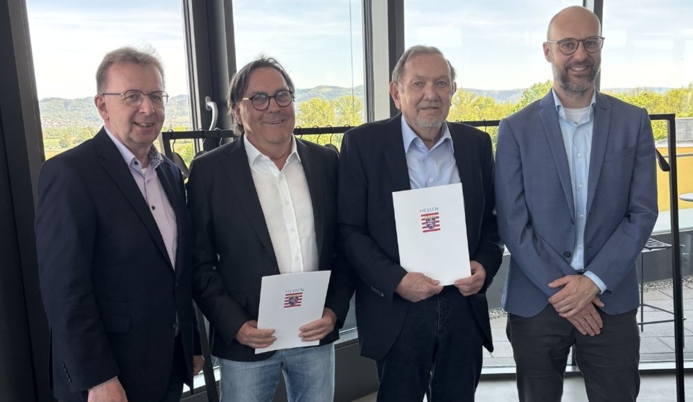 Vier Männer stehen nebeneinander in einem hellen Innenraum vor großen Fensterflächen mit Blick auf die grüne Landschaft vor den Hügeln der Bergstraße im Hintergrund. Sie schauen in die Kamera und stehen dicht beieinander, als Gruppenfoto. Zwei der Männer in der Mitte halten jeweils eine weiße Mappe mit dem hessischen Wappen und dem Wort „HESSEN“ darauf in den Händen. Der Raum wirkt modern, mit grauem Boden und einer Glasfront; rechts ist ein Stehtisch zu sehen. Von links nach rechts sind zu sehen: Bürgermeister Matthias Baaß, Josef Benz, Bernhard Seitz und Nils Bubeck. Alle vier tragen überwiegend dunkle oder gedeckte Kleidung, meist Sakko oder Blazer, kombiniert mit Hemd und langen Hosen. Die Stimmung wirkt ruhig und feierlich, passend zu einer Verabschiedung bzw. Ehrungssituation im Neuen Rathaus.