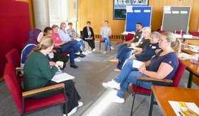 Eine Gruppe von elf Personen sitzt in einem hellen Raum auf roten Stühlen im Kreis. Einige halten Unterlagen in den Händen, während sie miteinander diskutieren. Im Hintergrund stehen Moderationstafeln. Die Arbeitsgruppe beschäftigt sich beim achten Runden Tisch „Im Alter gut versorgt in Viernheim“ mit der Unterstützung pflegebedürftiger Menschen mit Migrationsgeschichte und ihrer Angehörigen.