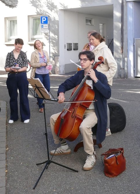 Ein junger Mann sitzt im Freien und spielt Cello. Vor ihm steht ein Notenständer. Der Musiker ist ein Familienmitglied von Frieda Horn. Im Hintergrund stehen mehrere Personen und verfolgen die musikalische Darbietung.