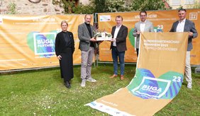 Stellen die Partnerschaft bei der BUGA 23 im Rahmen einer Pressekonferenz offiziell vor Fünf Personen stehen vor Transparenten mit Blumenkasten und Hinweistransparent