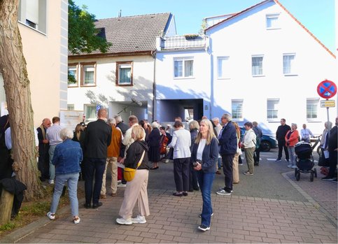 ine größere Gruppe von Menschen hat sich bei sonnigem Wetter auf einer Straße vor Wohnhäusern versammelt. Viele stehen in kleinen Gruppen zusammen.