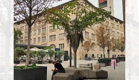 Öffentlicher Platz in Stuttgart mit einer mobilen Kühlinsel. Die Konstruktion ist mit Pflanzen bewachsen und überspannt mehrere Sitzgelegenheiten. Bäume und weitere Pflanzkübel ergänzen den begrünten Aufenthaltsbereich vor einem großen Gebäude.
