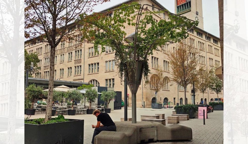 Öffentlicher Platz in Stuttgart mit einer mobilen Kühlinsel. Die Konstruktion ist mit Pflanzen bewachsen und überspannt mehrere Sitzgelegenheiten. Bäume und weitere Pflanzkübel ergänzen den begrünten Aufenthaltsbereich vor einem großen Gebäude.