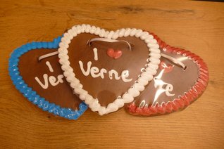 Drei herzförmige Lebkuchenherzen liegen leicht überlappend auf einem Holztisch. Sie sind mit blauem, weißem und rotem Zuckerguss umrandet. Auf jedem Herz steht in weißer Zuckerschrift „I ❤️ Verne“, wobei das Herzsymbol rot gestaltet ist. Das mittlere Lebkuchenherz mit weißem Rand liegt im Vordergrund, die beiden anderen sind teilweise verdeckt. Alle drei Herzen sind in durchsichtige Folie verpackt und mit weißen Bändern zum Umhängen versehen.