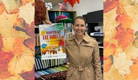 Renate Müller steht lächelnd in ihrer Nähgalerie und hält ein Plakat zur Aktion „Bunter Herbst“ in die Kamera. Im Geschäft sind herbstliche Stoffe und Dekorationen zu sehen.