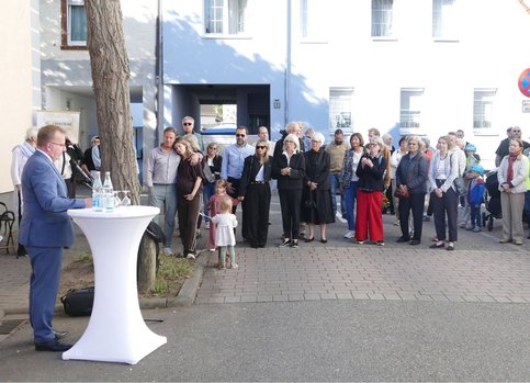 Bürgermeister Matthias Baaß steht links an einem Stehtisch und spricht über ein Mikrofon zu einer größeren Gruppe von Menschen. Die Zuhörenden haben sich im Freien vor mehreren Gebäuden versammelt. Unter ihnen sind Erwachsene und einige Kinder.