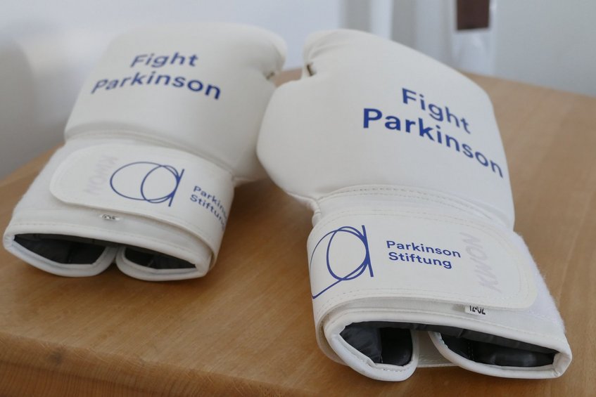 Zwei weiße Boxhandschuhe liegen nebeneinander auf einem hellen Holztisch. Auf beiden Handschuhen steht in blauer Schrift „Fight Parkinson“. Die breiten Klettverschlüsse an den Handgelenken tragen das Logo und den Schriftzug der Parkinson Stiftung. Der rechte Boxhandschuh liegt etwas weiter vorne. Der Hintergrund ist unscharf.