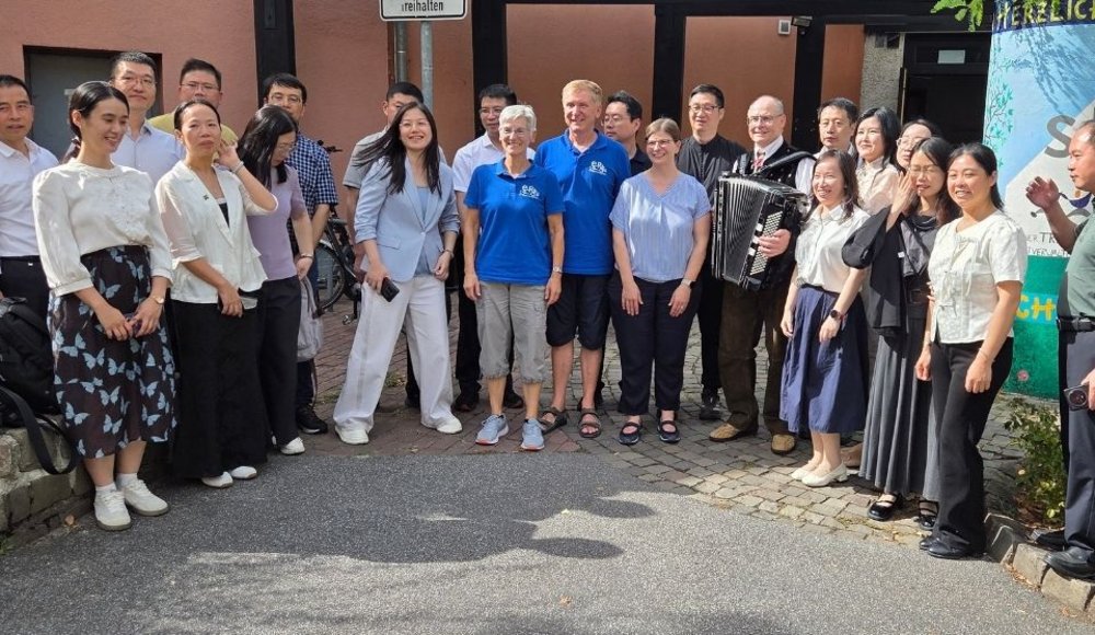 Eine größere Gruppe von Chinessinnen und Chinesen steht bei sonnigem Wetter vor dem Gebäude der Selbstverwalteten Begegnungsstätte 55+ für ein gemeinsames Gruppenfoto. In der Mitte hält ein Mann ein Akkordeon.