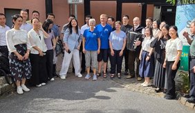 Eine größere Gruppe von Chinessinnen und Chinesen steht bei sonnigem Wetter vor dem Gebäude der Selbstverwalteten Begegnungsstätte 55+ für ein gemeinsames Gruppenfoto. In der Mitte hält ein Mann ein Akkordeon.