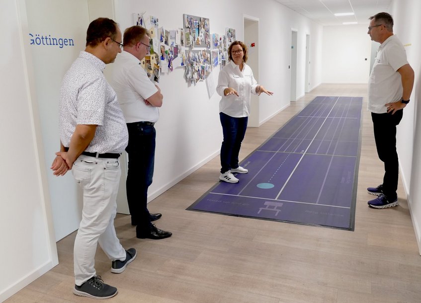In einem hellen Flur einer Praxis für Neurorehabilitation erklärt eine Therapeutin drei Männern eine Trainingsfläche zur Gang- und Gleichgewichtsschulung. Die lange, violette Bodenmatte ist mit Linien und Markierungen versehen. Die Therapeutin steht auf der Matte und gestikuliert mit beiden Händen, während die Männer ihr zuhören. An der Wand hängen zahlreiche Fotos.