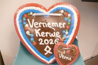 Zwei herzförmige Lebkuchenherzen liegen auf einem türkisfarbenen Tisch. Das große Herz ist mit einem rot-weiß-blauen Zuckergussrand und kleinen bunten Blumen verziert. In der Mitte steht in weißer Zuckerschrift „Vernemer Kerwe 2026“, darunter ist das Viernheimer Stadtwappen als Symbol dargestellt. Vor dem großen Herz steht rechts ein kleineres Lebkuchenherz mit rotem Zuckergussrand und der Aufschrift „I ❤️ Verne“. Beide Herzen sind in durchsichtige Folie verpackt und mit weißen Bändern zum Umhängen versehen.
