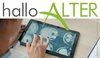 Eine Informationsgrafik mit dem Schriftzug „hallo-ALTER“ im oberen Bereich. Das Wort „hallo“ ist grau, „ALTER“ grün gestaltet. Darunter ist eine Person von hinten zu sehen, die mit den Fingern einen Tablet-Bildschirm bedient. Auf dem Bildschirm sind mehrere medizinische Schnittbilder eines menschlichen Gehirns zu erkennen. Das Tablet liegt auf einem hellen Holztisch. Am rechten Bildrand befindet sich ein schmaler Hinweis auf die Bildquelle und die Gestaltung durch die Stadt Viernheim.