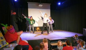 Auf einer kleinen Bühne tanzen fünf Mädchen und zeigen eine Choreografie mit ausgestreckten Armen. Im Vordergrund sitzen Kinder auf dem Boden und schauen zu, einige klatschen. Links sitzt eine verkleidete Person und applaudiert. Hinter den Tänzerinnen ist eine Leinwand aufgebaut, die Bühne ist von dunklen Vorhängen umgeben.