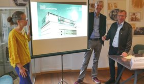 Bürgermeister Matthias Baaß (rechts), Dr. Harald Stockert und Elke Leinenweber präsentieren das MARCHIVUM, das nun die historischen Bestände des Viernheimer Stadtarchivs verwahrt. ©Stadt Viernheim Drei Personen stehen um Leinwand, auf welche ein Bild mit dem Gebäude Marchivum projeziert wird.