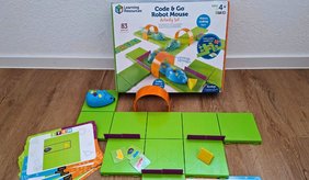 Aufgebautes Lernspiel mit einer blauen programmierbaren Robotermaus. Grüne Bodenplatten bilden einen Parcours mit kleinen Hindernissen und einem gelben Käse als Ziel. Daneben liegen Aufgabenkarten und Richtungskarten. Im Hintergrund steht die Verpackung des Lernspiels „Code & Go Robot Mouse“.