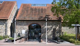 Außenansicht des Eingangs der Stadtbibliothek Viernheim in einem historischen Steingebäude mit rotem Ziegeldach. Vor dem Eingang befinden sich Sitzbänke, Bäume und eine Skulptur.