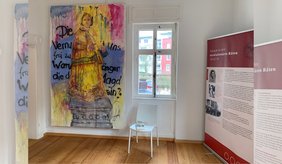 Ein heller Museumsraum mit Holzfußboden und weiß gestrichenen Wänden. Links hängt ein großes, buntes Gemälde: Es zeigt eine Frau in historischer Kleidung, die auf einem Sockel steht. Die Farben verlaufen ineinander, vor allem Gelb, Blau und Rot, und wirken teilweise wie herablaufende Farbtropfen. Über das Bild sind schwarze, handschriftliche Worte gesprüht, darunter Fragmente wie „Die Vernunft…“ und „frei zu sein“. In der Mitte befindet sich ein Fenster, durch das man Häuser und etwas Grün im Außenbereich sehen kann. Davor steht ein schlichter weißer Stuhl, auf dem eine kleine Broschüre liegt. Rechts steht eine große rote Informationstafel mit der Überschrift „Frauen in den revolutionären Räten 1918/19“. Darauf sind Texte und historische Fotos zu sehen, die sich mit der Rolle von Frauen in dieser Zeit beschäftigen.