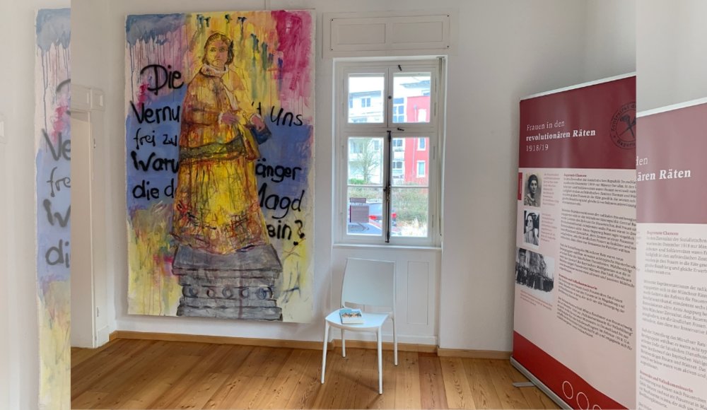 Ein heller Museumsraum mit Holzfußboden und weiß gestrichenen Wänden. Links hängt ein großes, buntes Gemälde: Es zeigt eine Frau in historischer Kleidung, die auf einem Sockel steht. Die Farben verlaufen ineinander, vor allem Gelb, Blau und Rot, und wirken teilweise wie herablaufende Farbtropfen. Über das Bild sind schwarze, handschriftliche Worte gesprüht, darunter Fragmente wie „Die Vernunft…“ und „frei zu sein“. In der Mitte befindet sich ein Fenster, durch das man Häuser und etwas Grün im Außenbereich sehen kann. Davor steht ein schlichter weißer Stuhl, auf dem eine kleine Broschüre liegt. Rechts steht eine große rote Informationstafel mit der Überschrift „Frauen in den revolutionären Räten 1918/19“. Darauf sind Texte und historische Fotos zu sehen, die sich mit der Rolle von Frauen in dieser Zeit beschäftigen.