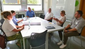 Sechs Personen sitzen in einem hellen Besprechungsraum an zusammengestellten weißen Tischen. Eine Frau gestikuliert während des Gesprächs, die anderen hören zu. Auf den Tischen liegen Unterlagen, Stifte und Wasserflaschen. Die Arbeitsgruppe beschäftigt sich beim achten Runden Tisch „Im Alter gut versorgt in Viernheim“ mit der gesundheitlichen Versorgung älterer Menschen mit Migrationsgeschichte.