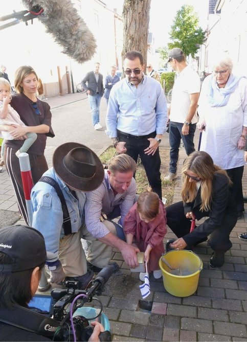 Mehrere Menschen verfolgen auf einem Gehweg die Verlegung von Stolpersteinen. Der Künstler und Initiator des Stolpersteinprojekts Gunter Demnig kniet links an der Pflasterstelle. Ein Mann und ein kleines Mädchen (Urenkelin von Frieda Horn) helfen beim Einsetzen eines Steins; das Mädchen hält eine kleine israelische Flagge. Weitere Personen stehen oder knien rundherum. Im Vordergrund filmt ein Kamerateam die Verlegung.