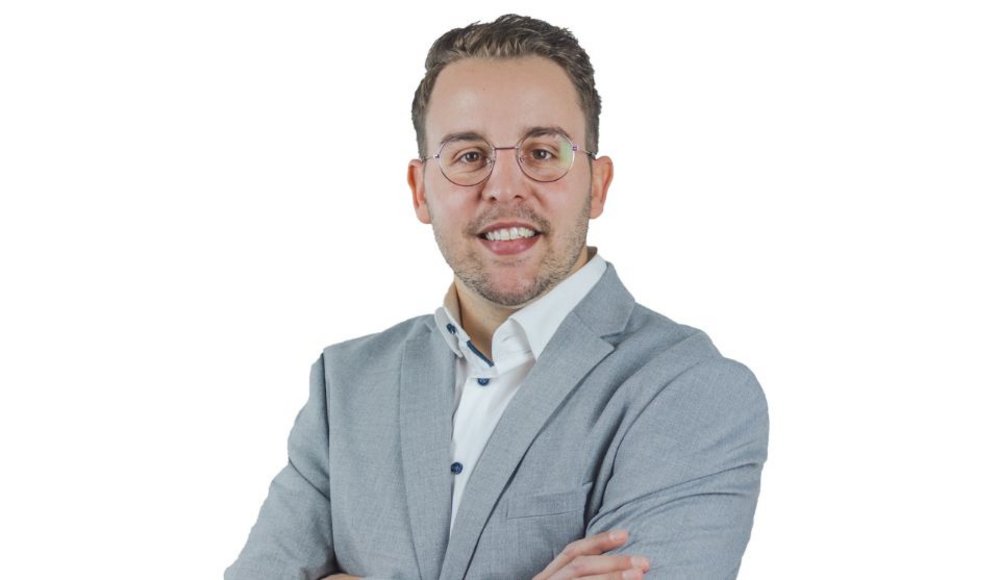 Porträt eines lächelnden Mannes, Jörg Scheidel, mit kurzen dunklen Haaren und runder Brille vor weißem Hintergrund. Er trägt ein weißes Hemd und ein hellgraues Sakko und hat die Arme vor der Brust verschränkt.
