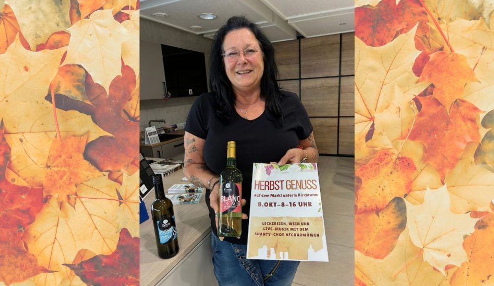 Eine lächelnde Frau mit langen dunklen Haaren steht in einem Geschäft und hält eine Weinflasche sowie ein Plakat in den Händen. Das Plakat kündigt die Veranstaltung „Herbst Genuss“ am 8. Oktober von 8 bis 16 Uhr an. Neben ihr steht eine weitere Weinflasche. Das Foto ist auf beiden Seiten mit einem dekorativen Rahmen aus gelb-orangefarbenem Herbstlaub versehen.