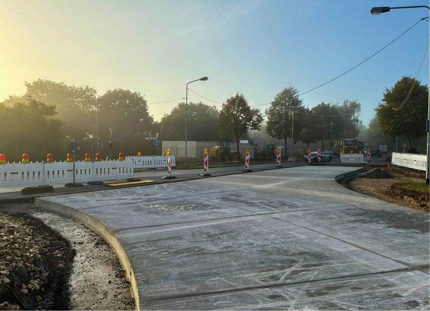 Eine Straßenbaustelle an einem Kreisverkehr bei Lampertheim-Hüttenfeld. Im Vordergrund ist eine neu betonierte, breite Fahrbahn zu sehen, die in einer Kurve nach rechts verläuft. Weiße Baustellenabsperrungen und rot-weiße Warnbaken mit gelben Warnleuchten sichern den Baustellenbereich. Im Hintergrund stehen mehrere Baufahrzeuge, darunter eine Walze und ein Lastwagen. Entlang der Straße wachsen hohe Bäume. Über der Baustelle liegt leichter Morgennebel, während von links die Sonne durch die Bäume scheint. Personen sind nicht zu sehen.