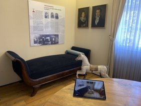 Tablet mit der Ausstellung und die Ausstellung - ein Schaukelpferd und ein Sofa - im Hintergund.