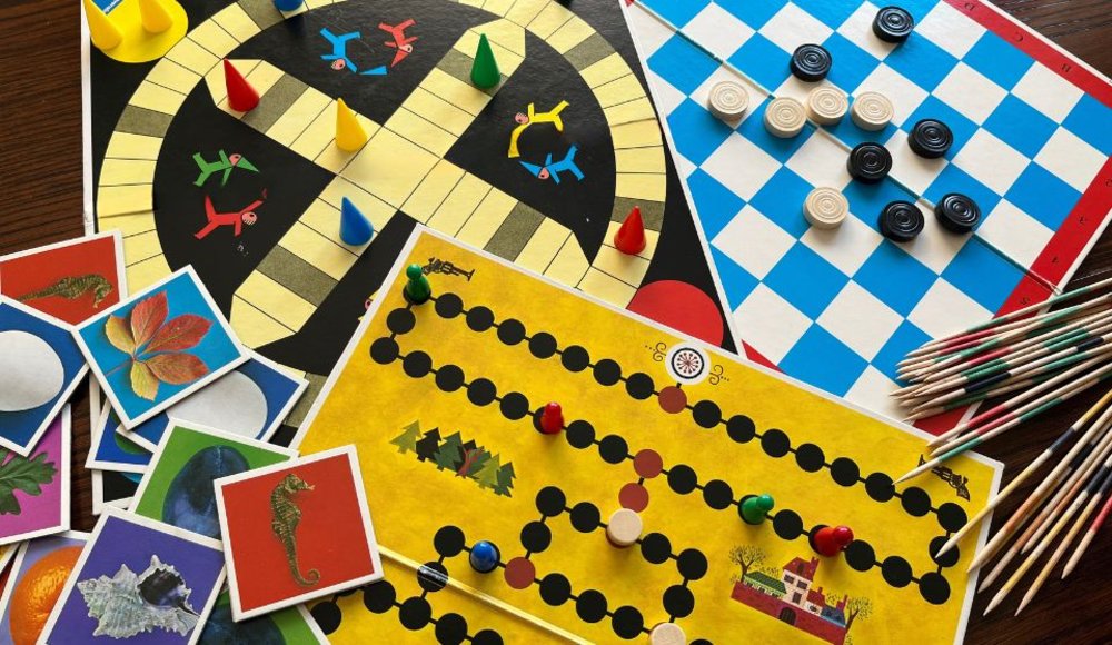 Mehrere Gesellschaftsspiele liegen auf einem Tisch. Zu sehen sind unter anderem bunte Spielfiguren auf Spielplänen, ein blau-weißes Damebrett mit schwarzen und weißen Spielsteinen sowie zahlreiche Mikado-Stäbchen. Links liegen Karten mit verschiedenen Bildmotiven. Das Foto zeigt eine vielfältige Auswahl an klassischen Brett-, Karten- und Geschicklichkeitsspielen.